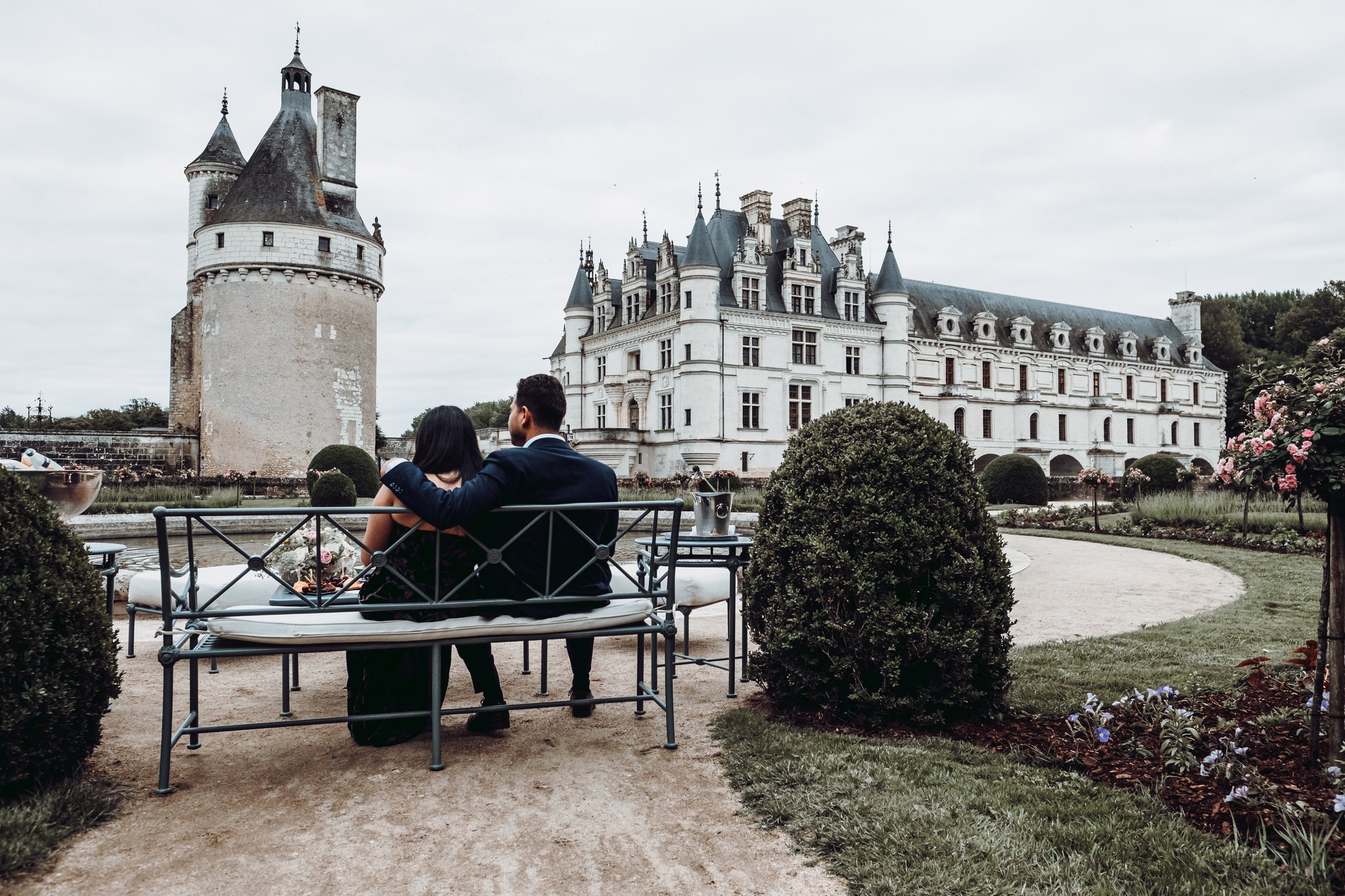 A couple on a garden bench facing the floodlit Château de Chenonceau, champagne chilling beside them